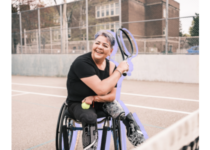 A woman with prosthetic legs sat in a wheelchair on a tennis court in an urban setting holding a tennis racket.