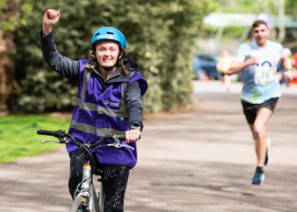 Woman riding a bike wearing a purple CSSC jacket followed by a runner in the background  at Battersea Park.