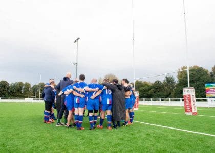 A mens rugby team stood on a rugby pitch in front of the goal with a purple CSSC standing banner in front of them