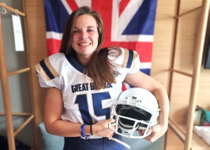 Steph smiling wearing her american football kit holding her helmet with a GB flag behind her.